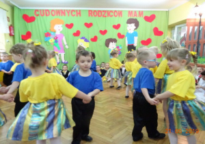 Dziewczynki ubrane w żółte podkoszulki i zielono-żółtto-niebieskie spódniczki oraz chłopcy w niebieskich podkoszulkach i czarnych spodniach wykonują dla rodziców taniec w parach.
